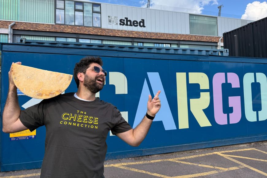 a man holding a big cheese wheel pointing at Cargo